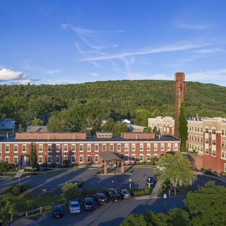 A historic brick factory building with a tall chimney, surrounded by greenery and a clear blue sky.