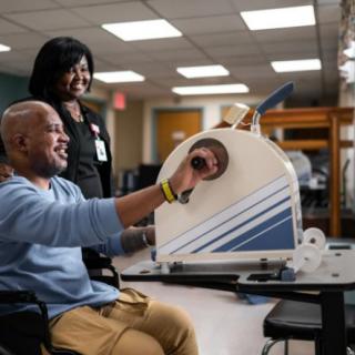 A man in a wheelchair uses therapy equipment while being assisted by a healthcare professional.