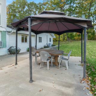 An outdoor patio area with a gazebo covering a wooden table and chairs set, surrounded by a grassy lawn.