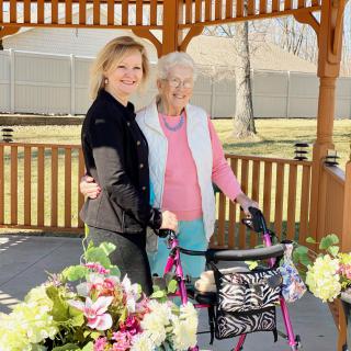 An elderly woman and a young woman standing together outdoors in a gazebo surrounded by flowers, with the elderly woman using a walker.