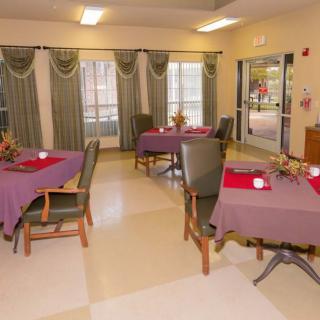 A cozy dining area with tables set for a meal, featuring purple tablecloths and natural light from large windows.