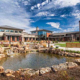 A serene outdoor scene featuring a pond surrounded by rocks, with buildings in the background under a partly cloudy sky.