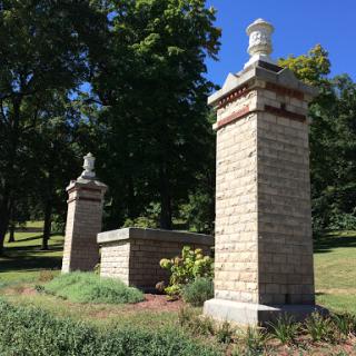 A historic stone monument with decorative pillars and a landscaped area, set against a backdrop of trees.
