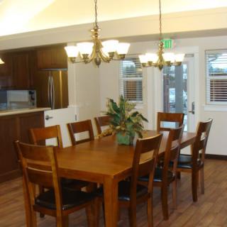 A spacious dining area with a wooden table and chairs, illuminated by a chandelier, connecting to a kitchen.