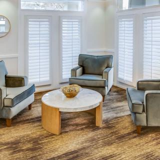 A cozy living room with three armchairs surrounding a round coffee table, featuring wooden flooring and window shutters.