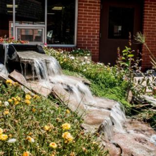 A small waterfall cascading through a garden with flowers in front of a brick building.
