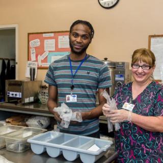 Two people working together in a kitchen, preparing meals and wearing uniforms.
