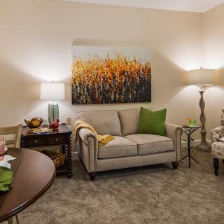 A cozy living room with a beige sofa, decorated with green pillows, flanked by lamps and a vibrant nature painting on the wall.