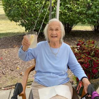 An elderly woman sitting outdoors, smiling and holding up a drink.