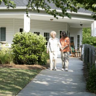An elderly person walking with a caregiver outside a house on a sunny day.