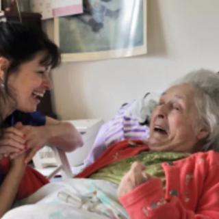 A caregiver shares a joyful moment with an elderly woman in bed.