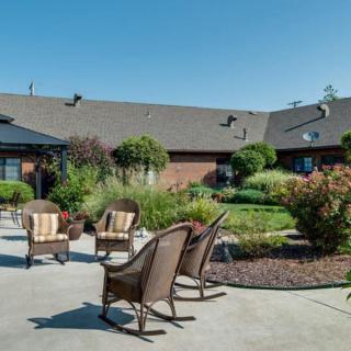 A relaxing outdoor patio with chairs surrounded by a landscaped garden and a brick building.