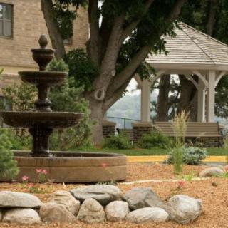 A peaceful garden scene with a fountain, gazebo, and bench surrounded by trees.