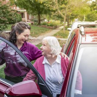 A caregiver assisting an elderly woman as she gets out of a car in a residential neighborhood.