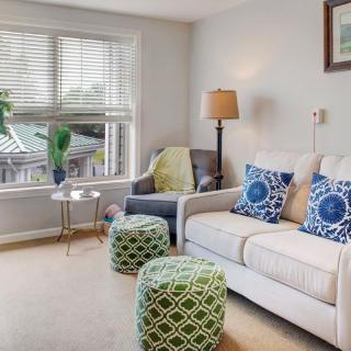 A cozy living room with a sofa and armchair adorned with colorful pillows, accompanied by a floor lamp and vibrant indoor plants near the window.