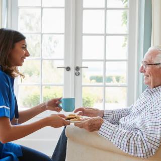 A caregiver offering tea and cookies to an elderly man during a home visit.