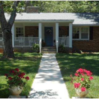 A quaint brick house with a porch and a walkway lined with flowers in a lush front yard.