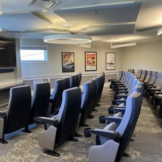 A small auditorium with rows of cushioned seats facing a projector screen.