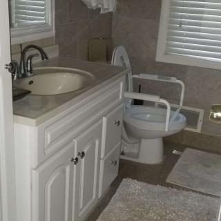 A bathroom featuring a sink with a cabinet, toilet with handrails, and two rugs on the floor.