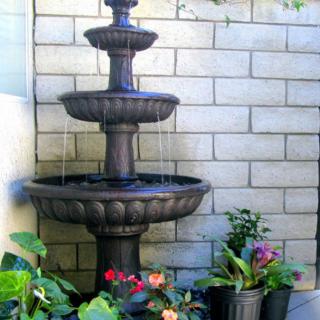 A three-tier water fountain surrounded by colorful potted plants, set against a brick wall.