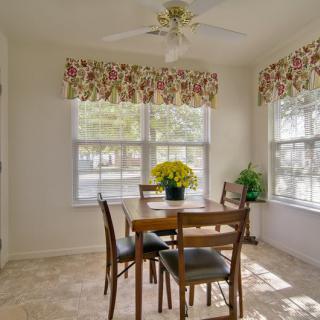 A bright dining area with natural light, featuring a wooden table and chairs surrounded by large windows with floral curtains.