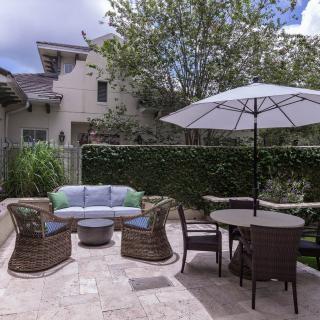 A cozy patio with a seating area, table, and umbrella next to a green hedge in a backyard.