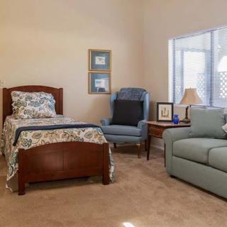 A cozy bedroom with a single bed, armchair, and sofa, featuring decorative elements and a window letting in natural light.