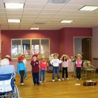 A group of children wearing hats perform indoors in front of an audience.