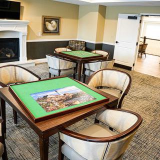 A cozy game room with tables set for puzzles, surrounded by comfortable chairs, a fireplace, and artwork on the walls.