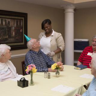 A group of senior citizens celebrating a birthday at a nursing home with party hats and flowers on the table.