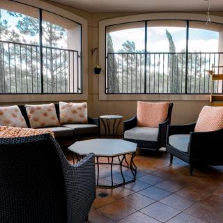 A cozy outdoor seating area with cushioned chairs and sofas positioned around small tables on a tiled floor, enclosed by large windows.