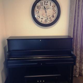 A black piano with a wall-mounted clock above in a cozy interior setting.