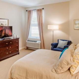 A cozy bedroom with a neatly made bed, a wooden dresser with a TV on top, and a comfortable armchair by the window.