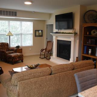 A cozy living room featuring a sofa, fireplace with a mounted TV, rocking chair, and bookshelf.