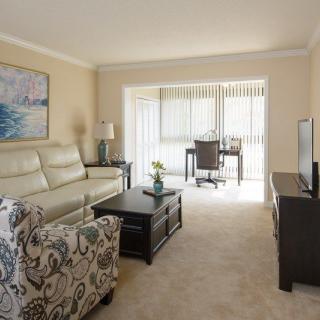 A cozy living room with a sofa, patterned armchair, and coffee table, featuring a desk near a window with blinds.