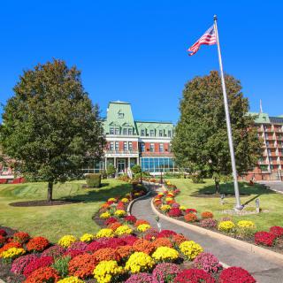 A beautifully landscaped garden with colorful flowers leading to a large building, accompanied by a flagpole and trees under a clear blue sky.