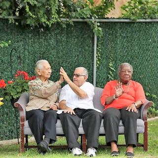 Three seniors sitting on a bench outdoors, enjoying a conversation in a garden setting.