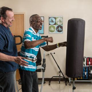 A senior citizen engages in a boxing exercise with the assistance of a caregiver, promoting fitness and rehabilitation.
