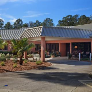 A single-story building with a landscaped entrance and parking area, featuring a palm tree and surrounding trees.
