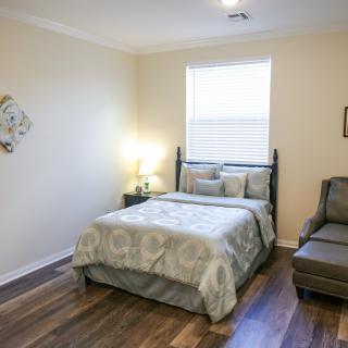 A cozy bedroom with a neatly made bed, an armchair, and a lamp beside the window.