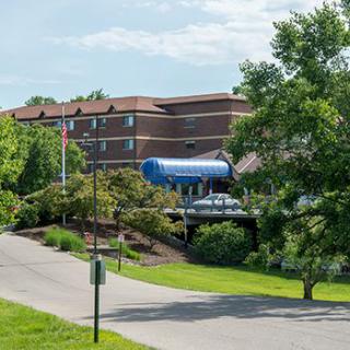 A building with a blue awning is surrounded by lush trees, with a road leading up to it and a flagpole visible in the foreground.