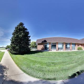 A brick building with a sloped roof adjacent to a large tree and a grassy area under a clear blue sky.