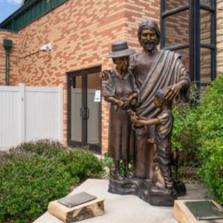 A statue of two figures stands in front of a brick building surrounded by greenery.