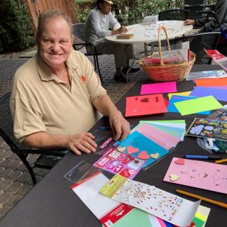 A man sitting at an outdoor table with colorful crafting supplies and smiling.
