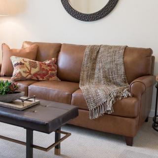 A stylish living room featuring a brown leather couch adorned with decorative pillows and a cozy throw blanket, accompanied by a modern coffee table.