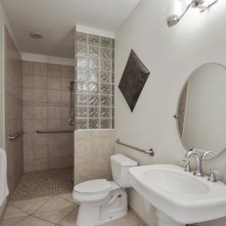 A modern bathroom featuring a walk-in shower, white toilet and sink, and an oval mirror above the sink. The space is accented with a glass block wall and tiled flooring.