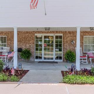 A brick building entrance with a porch featuring rocking chairs, an American flag, and decorative plants.