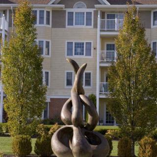 An abstract sculpture set in a landscaped area with trees and a residential building in the background.