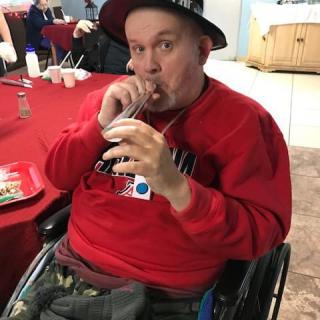 A man in a wheelchair wearing a hat and sipping a drink through a straw indoors.