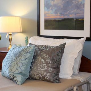 A cozy bedroom setup featuring decorative pillows, a bedside lamp, and a framed painting above the bed.